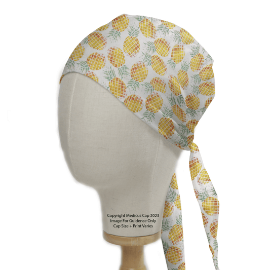 A mannequin head showcases the Tropical Pineapples Scrub Cap by Medicus Scrub Caps, adorned with vibrant yellow pineapples and lush green leaves on a crisp white background. This reusable cap features long ties, merging sustainability with stylish medical attire.