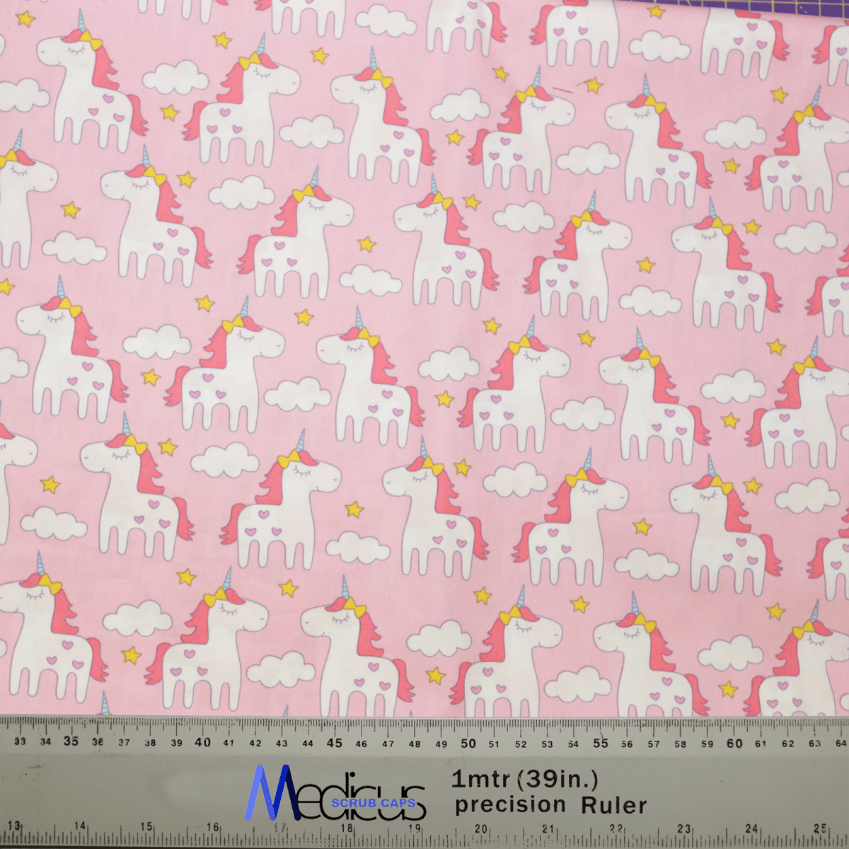 The Unicorn Cartoon Pink Scrub Cap by Medicus Scrub Caps showcases white unicorns with rainbow manes and tails, yellow-starred collars, on a pink background with white clouds and yellow stars. A ruler in centimeters and inches is included, making it ideal for crafting sustainable scrub caps.