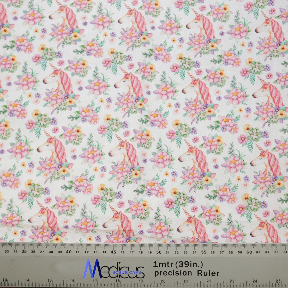 The Medicus Scrub Caps Unicorns Flower Power Scrub Cap features fabric with pink unicorns and colorful flowers on a white background, ideal for crafting. A transparent ruler marks centimeters and inches for precise cutting.