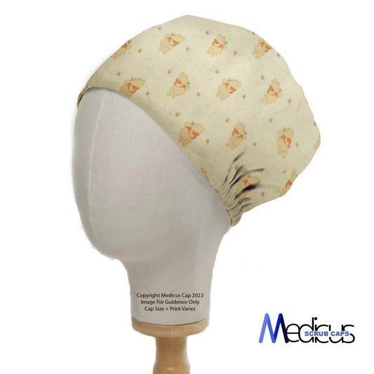 A mannequin head displays the Winnie The Pooh Honey + Bees Scrub Cap by Medicus Scrub Caps, featuring cartoon puppies and playful honey bees on a light-colored fabric. The caps soft material and elastic section ensure a snug fit, with the brands logo at the bottom right corner.