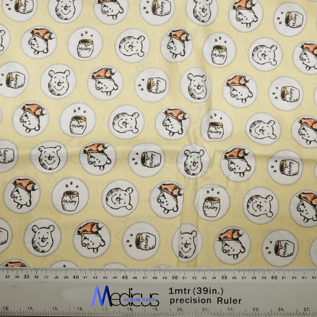 The Winnie the Pooh Honey Pots Scrub Cap by Medicus Scrub Caps features yellow fabric with cartoon bear faces and Hunny pots, alternating with orange hats and plain designs, perfect for healthcare professionals. A 1-meter precision ruler is placed along the fabric’s edge.