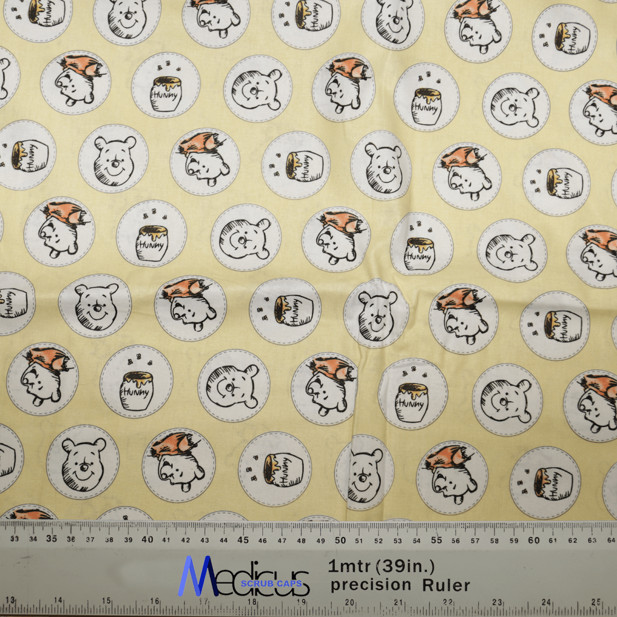The Winnie the Pooh Honey Pots Scrub Cap by Medicus Scrub Caps features yellow fabric with cartoon bear faces and Hunny pots, alternating with orange hats and plain designs, perfect for healthcare professionals. A 1-meter precision ruler is placed along the fabric’s edge.